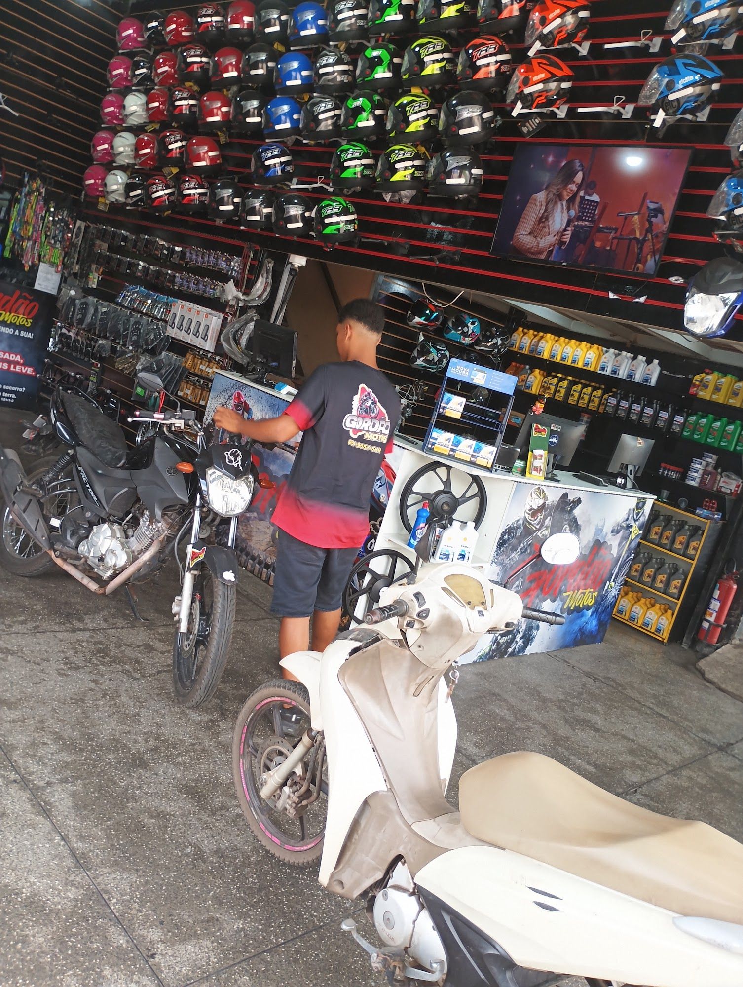 Atendente da Gordão Motos no balcão, com parede de capacetes e prateleiras de óleo ao fundo